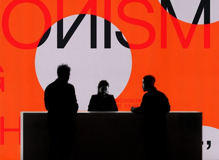 Three figures, backs to us, facing a lit wall of mirrored type